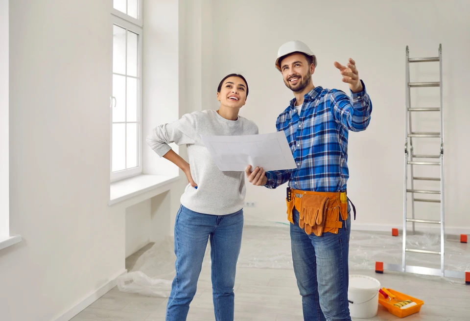 Superviseur de la rénovation ou constructeur à hardhat rencontre une jeune femme heureuse propriétaire et montre son processus de peinture et de décoration dans une nouvelle maison ou appartement vide, avec échelle et seau en arrière-plan