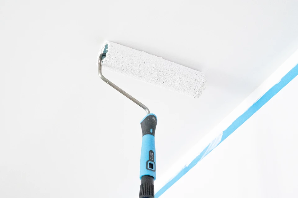 Un homme en t-shirt gris installe du papier peint blanc sur un mur intérieur, debout sur un escabeau télescopique en aluminium